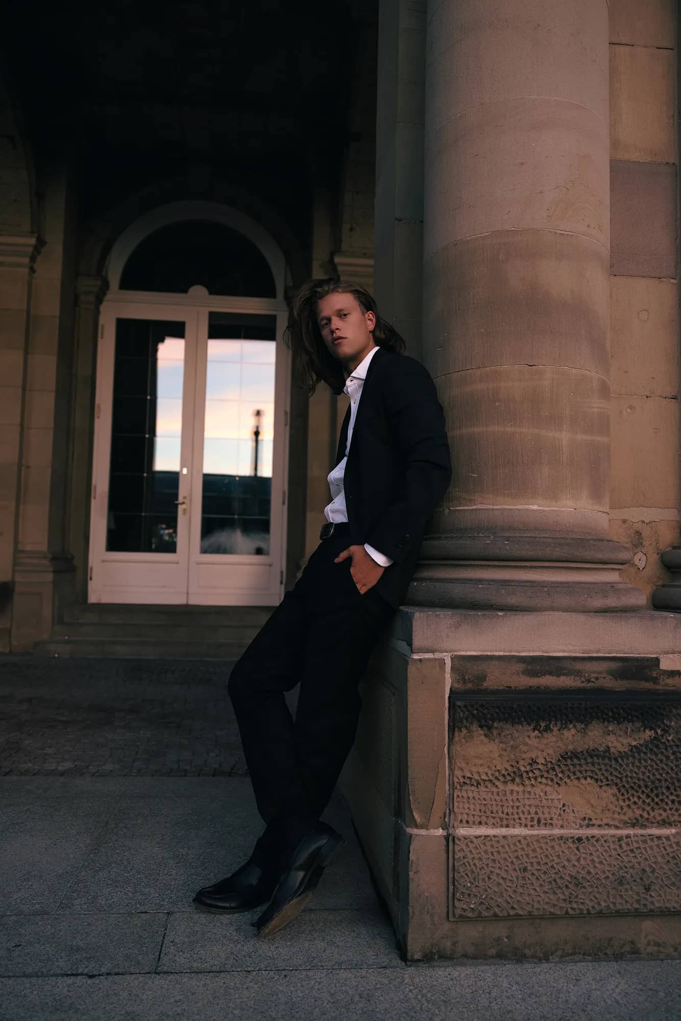 Ein eleganter Mann im Anzug lehnt sich an eine Säule und posiert vor einem Schloss.
