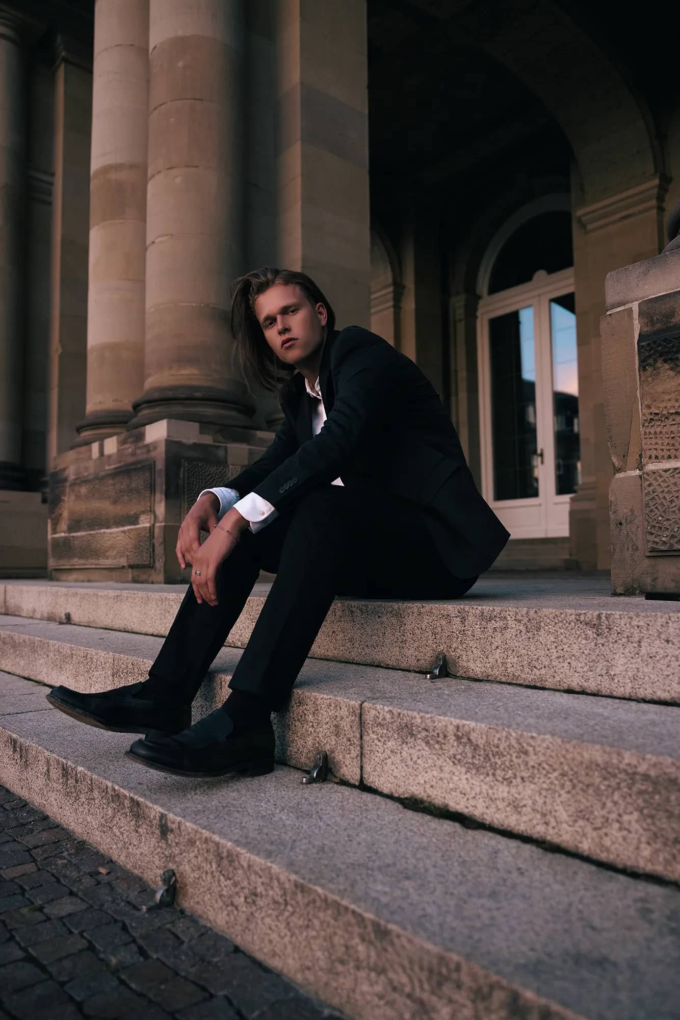 Ein eleganter Mann in Anzug sitzt auf einer Treppe vor einem Schloss und posiert stilvoll.